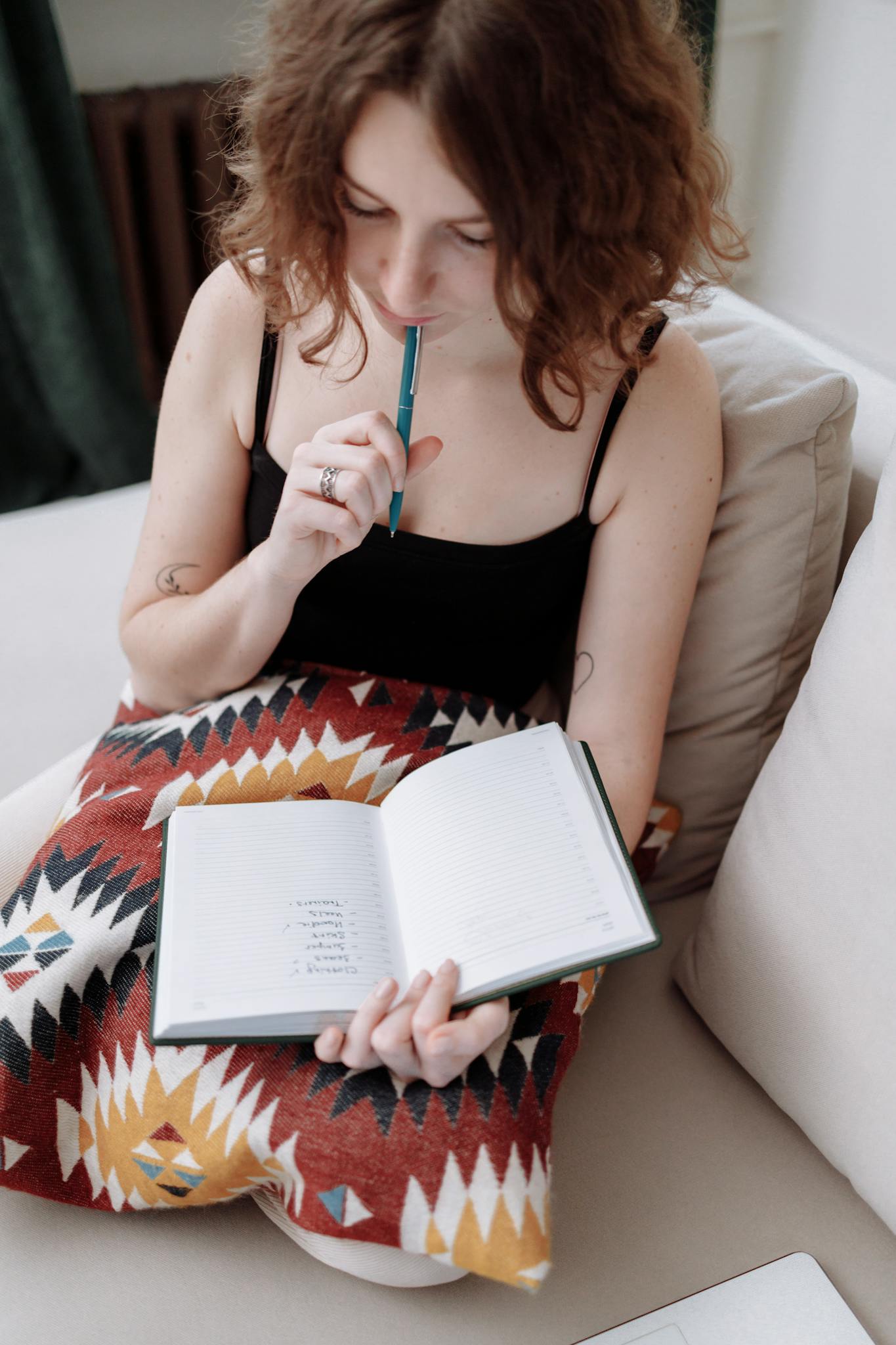 A woman sitting on a sofa, pondering while holding an open notebook and pen.